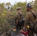 Two wildland firefighters are working in a brushy, partially burned landscape. The foreground firefighter is holding a drip torch (a red canister with an active flame), indicating they are conducting a prescribed burn or backfire operation. Both personnel are wearing standard wildland firefighting gear including hard hats, fire-resistant Nomex clothing, and are carrying tools and equipment.