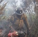 A wildland firefighter wearing a U.S. Forest Service hard hat and fire-resistant gear is working in a smoky, burning brush area. The firefighter is holding a drip torch, a device used to ignite controlled burns or manage fire lines. The environment is heavily obscured by smoke with visible flames and charred vegetation in the foreground.