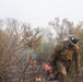 A wildland firefighter is working in dense, smoke-filled brush while actively combating a wildfire. The firefighter is holding a red drip torch or similar firefighting tool and is surrounded by burning and scorched vegetation. The scene depicts close-range ground-level fire suppression or controlled burn operations in rugged terrain.