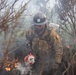 A U.S. Forest Service wildland firefighter is actively working in a smoky, burning landscape surrounded by charred and burning brush. The firefighter is carrying a drip torch, a tool used to ignite or manage controlled burns and fire lines. The scene depicts intense, close-quarters wildfire suppression or prescribed burn operations.