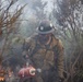 A U.S. Forest Service wildland firefighter is working in a smoky, burning landscape surrounded by charred and partially burned vegetation. The firefighter is carrying a drip torch, a tool used to ignite controlled burns or manage fire lines. The individual is wearing standard wildland firefighting gear including a dirty, soot-covered Nomex suit and a hard hat marked with 'U.S.' insignia.