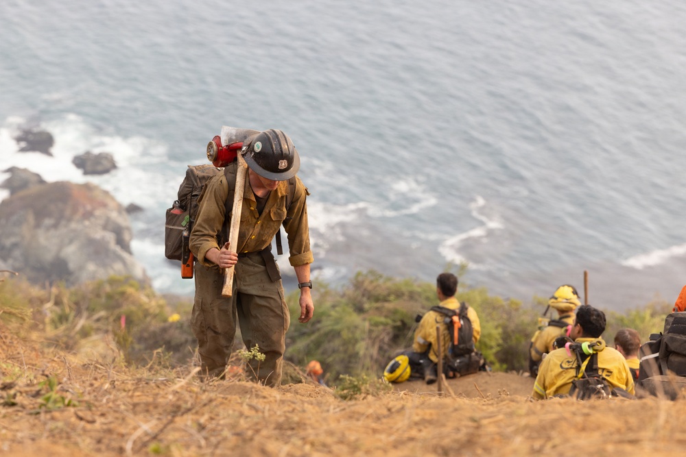 A group of wildland firefighters are working on a steep coastal hillside with the ocean visible in the background. The foreground firefighter is wearing standard wildland fire gear including a hard hat, Nomex suit, and pack, and is carrying a hand tool. Additional crew members in yellow Nomex shirts are resting or sitting on the slope to the right.