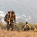 A group of wildland firefighters are working on a steep coastal hillside with the ocean visible in the background. The foreground firefighter is wearing standard wildland fire gear including a hard hat, Nomex suit, and pack, and is carrying a hand tool. Additional crew members in yellow Nomex shirts are resting or sitting on the slope to the right.
