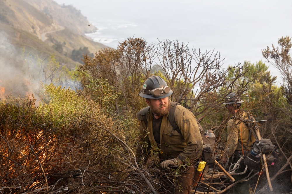 A wildland firefighter wearing a gray hard hat with goggles and dirty protective gear works on a steep coastal hillside amid active fire and smoke. At least one additional firefighter is visible in the background. The scene appears to be a wildfire suppression operation along a rugged coastal terrain, likely in California based on the landscape.