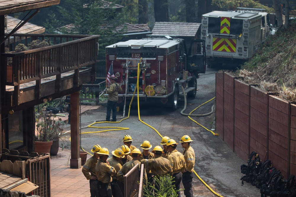A group of approximately 10-12 wildland firefighters in yellow helmets and tan nomex gear are gathered in a briefing or debriefing near a residential structure. A red fire engine marked 'E-302' is positioned in the background with yellow hoses deployed, alongside a second larger utility vehicle with yellow chevron markings. The scene shows signs of fire damage to surrounding vegetation and structures.