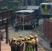 A group of approximately 10-12 wildland firefighters in yellow helmets and tan nomex gear are gathered in a briefing or debriefing near a residential structure. A red fire engine marked 'E-302' is positioned in the background with yellow hoses deployed, alongside a second larger utility vehicle with yellow chevron markings. The scene shows signs of fire damage to surrounding vegetation and structures.