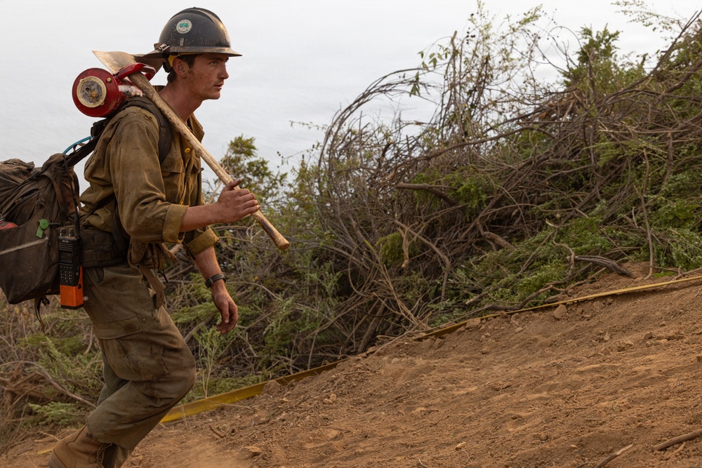A wildland firefighter is walking along a dirt firebreak or hand line on a hillside. He is carrying hand tools and wearing full personal protective equipment, appearing dirty and worn from active fire suppression work. The surrounding vegetation shows signs of disturbance consistent with firefighting operations.