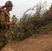 A wildland firefighter is walking along a dirt firebreak or hand line on a hillside. He is carrying hand tools and wearing full personal protective equipment, appearing dirty and worn from active fire suppression work. The surrounding vegetation shows signs of disturbance consistent with firefighting operations.