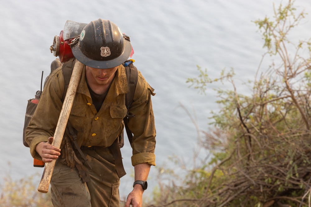 A U.S. Forest Service wildland firefighter is captured climbing uphill near a body of water, visibly fatigued and heavily equipped. He is wearing standard wildland firefighting gear including a dirty Nomex shirt, hard hat with a U.S. Forest Service shield emblem, and is carrying a hand tool (likely a Pulaski or similar firefighting tool) over his shoulder. His gear shows significant wear and dirt, indicating active field deployment.