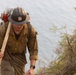 A U.S. Forest Service wildland firefighter is captured climbing uphill near a body of water, visibly fatigued and heavily equipped. He is wearing standard wildland firefighting gear including a dirty Nomex shirt, hard hat with a U.S. Forest Service shield emblem, and is carrying a hand tool (likely a Pulaski or similar firefighting tool) over his shoulder. His gear shows significant wear and dirt, indicating active field deployment.