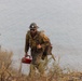 A wildland firefighter is climbing up a steep, fire-affected hillside near a body of water. The worker is heavily equipped and visibly dirty, suggesting active or recent firefighting operations. They are carrying a drip torch, a tool used to intentionally set controlled burns.