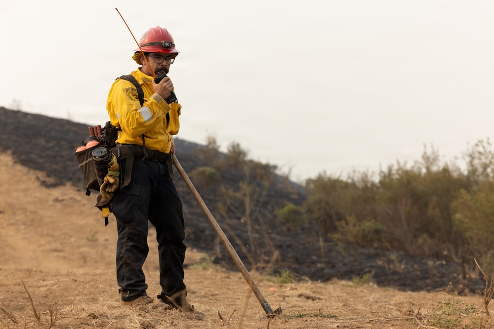 A wildland firefighter stands on a hillside communicating via handheld radio while holding a hand tool, likely a Pulaski or similar firefighting implement. The background shows a scorched hillside with blackened earth, indicating a recent fire burn area. The firefighter is equipped with standard wildland fire personal protective equipment.