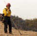 A wildland firefighter stands on a hillside communicating via handheld radio while holding a hand tool, likely a Pulaski or similar firefighting implement. The background shows a scorched hillside with blackened earth, indicating a recent fire burn area. The firefighter is equipped with standard wildland fire personal protective equipment.