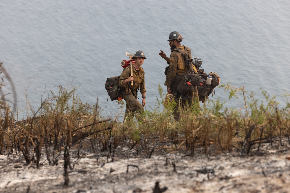 Two wildland firefighters are walking through a recently burned area near a body of water. They are fully equipped with gear, tools, and heavy packs, suggesting they are either arriving at or departing from a fire suppression operation. The foreground shows charred vegetation and ash, indicating recent fire activity in the area.