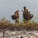 Two wildland firefighters are walking through a recently burned area near a body of water. They are fully equipped with gear, tools, and heavy packs, suggesting they are either arriving at or departing from a fire suppression operation. The foreground shows charred vegetation and ash, indicating recent fire activity in the area.