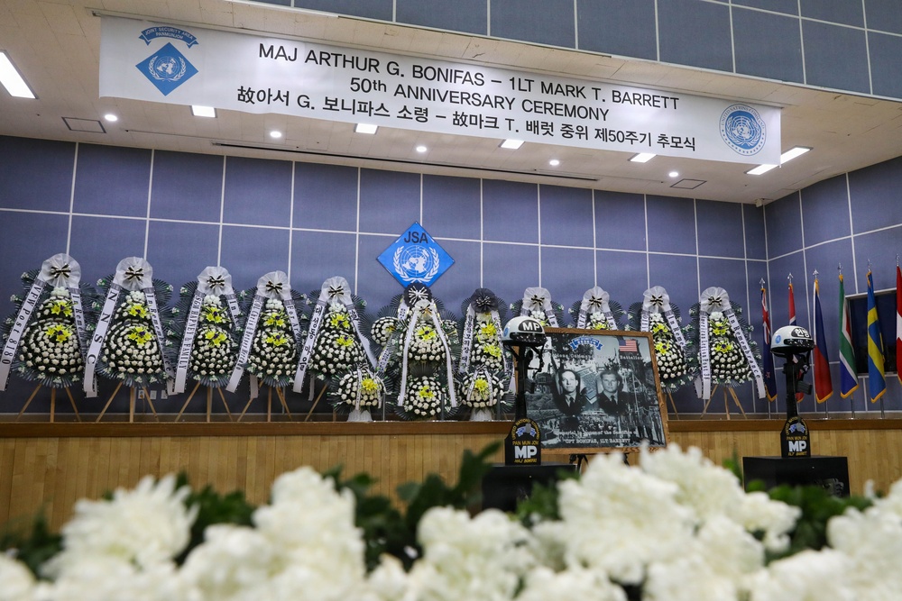 This image shows a memorial ceremony stage commemorating the 50th anniversary of the deaths of MAJ Arthur G. Bonifas and 1LT Mark T. Barrett, as indicated by the banner overhead. The stage is decorated with numerous traditional Korean funeral flower wreaths (근조화환) arranged in a row, along with military MP helmets, a framed photograph, and international flags. The ceremony appears to be held at the Joint Security Area (JSA), as indicated by the JSA emblem displayed on the backdrop.