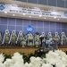 This image shows a memorial ceremony stage commemorating the 50th anniversary of the deaths of MAJ Arthur G. Bonifas and 1LT Mark T. Barrett, as indicated by the banner overhead. The stage is decorated with numerous traditional Korean funeral flower wreaths (근조화환) arranged in a row, along with military MP helmets, a framed photograph, and international flags. The ceremony appears to be held at the Joint Security Area (JSA), as indicated by the JSA emblem displayed on the backdrop.