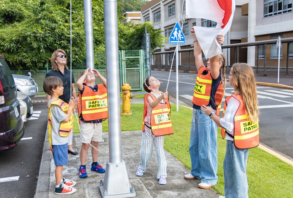 A group of children wearing orange 'Safety Patrol' vests are raising a flag on a flagpole near a school, supervised by an adult woman. The scene appears to take place outside a school in Japan, evidenced by a Japanese school zone crossing sign and surrounding architecture. The children are actively participating in what appears to be a flag-raising ceremony or safety patrol duty.