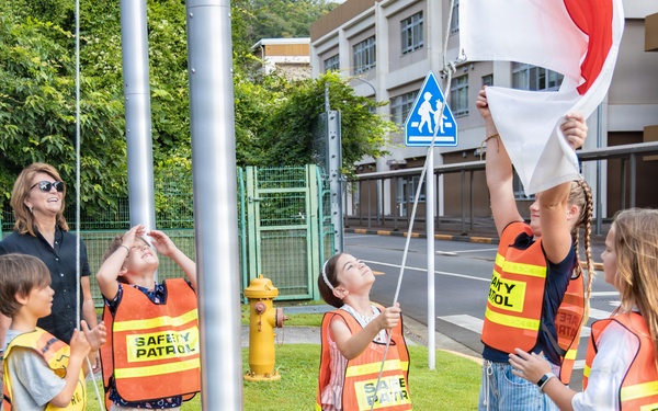A group of children wearing orange 'Safety Patrol' vests are raising a flag on a flagpole near a school, supervised by an adult woman. The scene appears to take place outside a school in Japan, evidenced by a Japanese school zone crossing sign and surrounding architecture. The children are actively participating in what appears to be a flag-raising ceremony or safety patrol duty.