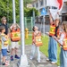 A group of children wearing orange 'Safety Patrol' vests are raising a flag on a flagpole near a school, supervised by an adult woman. The scene appears to take place outside a school in Japan, evidenced by a Japanese school zone crossing sign and surrounding architecture. The children are actively participating in what appears to be a flag-raising ceremony or safety patrol duty.