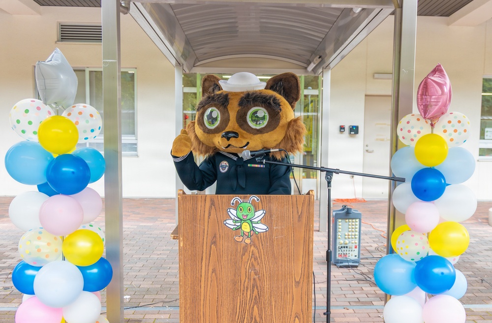 A person wearing a large animal mascot costume (resembling a raccoon or tanuki) dressed in a dark naval/military uniform with a white sailor cap stands at a wooden podium with a microphone. The event appears to be a celebratory ceremony, flanked by colorful balloon columns on both sides. A cartoon dragonfly or bee character logo is visible on the front of the podium.