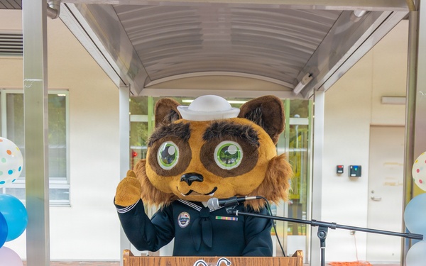 A person wearing a large animal mascot costume (resembling a raccoon or tanuki) dressed in a dark naval/military uniform with a white sailor cap stands at a wooden podium with a microphone. The event appears to be a celebratory ceremony, flanked by colorful balloon columns on both sides. A cartoon dragonfly or bee character logo is visible on the front of the podium.