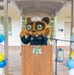 A person wearing a large animal mascot costume (resembling a raccoon or tanuki) dressed in a dark naval/military uniform with a white sailor cap stands at a wooden podium with a microphone. The event appears to be a celebratory ceremony, flanked by colorful balloon columns on both sides. A cartoon dragonfly or bee character logo is visible on the front of the podium.