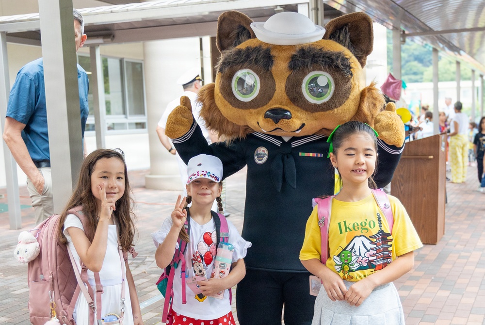 Three young girls pose with a Navy-themed mascot costume character wearing a sailor uniform and white sailor cap. The setting appears to be a school or community event, likely at a U.S. Navy installation in Japan based on the 'Ikego' text visible on one girl's yellow shirt. The children are wearing backpacks, suggesting a back-to-school or welcome event.