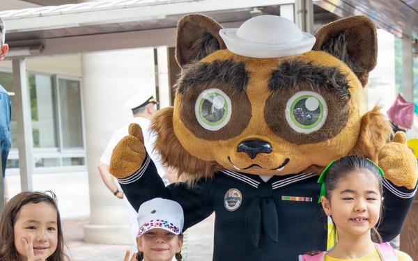 Three young girls pose with a Navy-themed mascot costume character wearing a sailor uniform and white sailor cap. The setting appears to be a school or community event, likely at a U.S. Navy installation in Japan based on the 'Ikego' text visible on one girl's yellow shirt. The children are wearing backpacks, suggesting a back-to-school or welcome event.