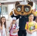 Three young girls pose with a Navy-themed mascot costume character wearing a sailor uniform and white sailor cap. The setting appears to be a school or community event, likely at a U.S. Navy installation in Japan based on the 'Ikego' text visible on one girl's yellow shirt. The children are wearing backpacks, suggesting a back-to-school or welcome event.