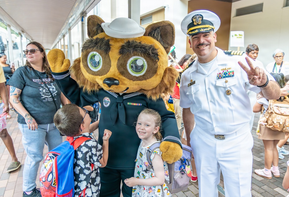 A U.S. Navy officer in white dress uniform poses with a Navy mascot character (wearing a sailor uniform) and two young children at what appears to be a school or community event. The officer is making a peace sign while the mascot waves, creating a cheerful, celebratory atmosphere. The setting appears to be an outdoor covered walkway with a crowd of adults and children in the background.