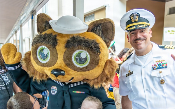 A U.S. Navy officer in white dress uniform poses with a Navy mascot character (wearing a sailor uniform) and two young children at what appears to be a school or community event. The officer is making a peace sign while the mascot waves, creating a cheerful, celebratory atmosphere. The setting appears to be an outdoor covered walkway with a crowd of adults and children in the background.