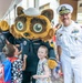 A U.S. Navy officer in white dress uniform poses with a Navy mascot character (wearing a sailor uniform) and two young children at what appears to be a school or community event. The officer is making a peace sign while the mascot waves, creating a cheerful, celebratory atmosphere. The setting appears to be an outdoor covered walkway with a crowd of adults and children in the background.