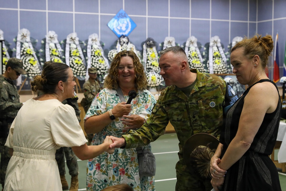 A military ceremony or reception is taking place in what appears to be an indoor venue, likely in South Korea based on the JSA (Joint Security Area) and United Nations insignia visible in the background. A soldier in Australian Defence Force camouflage uniform is shaking hands with a civilian woman while another woman holds a microphone nearby. Floral wreaths typical of Korean ceremonial traditions line the back wall.