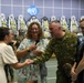 A military ceremony or reception is taking place in what appears to be an indoor venue, likely in South Korea based on the JSA (Joint Security Area) and United Nations insignia visible in the background. A soldier in Australian Defence Force camouflage uniform is shaking hands with a civilian woman while another woman holds a microphone nearby. Floral wreaths typical of Korean ceremonial traditions line the back wall.