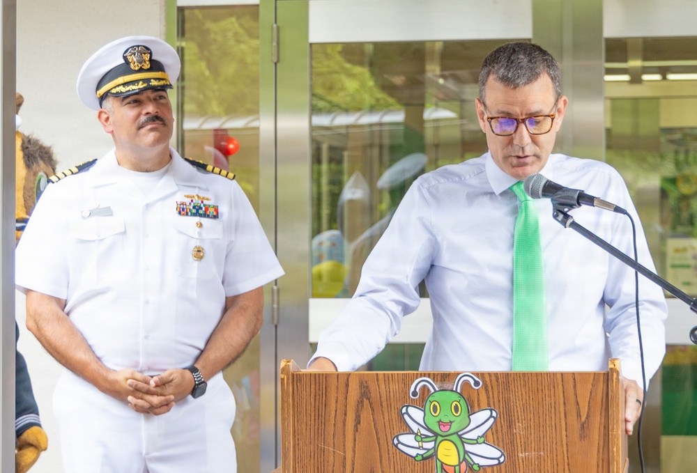 Two men are standing at what appears to be an outdoor or semi-outdoor public event. One man in a white U.S. Navy officer's dress uniform with rank insignia and ribbons stands to the left, while a civilian man in a light blue shirt and green tie speaks at a wooden podium equipped with a microphone. The podium features a cartoon bee mascot logo, suggesting a school or children's organization setting.