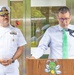 Two men are standing at what appears to be an outdoor or semi-outdoor public event. One man in a white U.S. Navy officer's dress uniform with rank insignia and ribbons stands to the left, while a civilian man in a light blue shirt and green tie speaks at a wooden podium equipped with a microphone. The podium features a cartoon bee mascot logo, suggesting a school or children's organization setting.