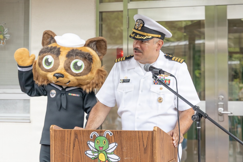 A U.S. Navy officer in white dress uniform with gold rank insignia and service ribbons speaks at a wooden podium equipped with a microphone. Behind him stands a costumed mascot character — a cartoon raccoon/cat figure wearing a Navy enlisted uniform with a white sailor cap and service ribbons. The podium features a green bee mascot logo, suggesting a school or youth-oriented event setting.