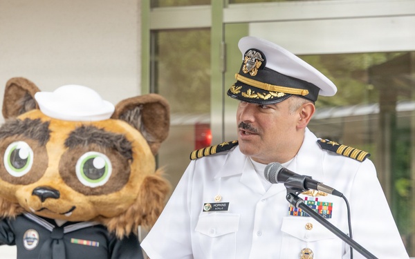 A U.S. Navy officer in white dress uniform with gold rank insignia and service ribbons speaks at a wooden podium equipped with a microphone. Behind him stands a costumed mascot character — a cartoon raccoon/cat figure wearing a Navy enlisted uniform with a white sailor cap and service ribbons. The podium features a green bee mascot logo, suggesting a school or youth-oriented event setting.