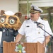 A U.S. Navy officer in white dress uniform with gold rank insignia and service ribbons speaks at a wooden podium equipped with a microphone. Behind him stands a costumed mascot character — a cartoon raccoon/cat figure wearing a Navy enlisted uniform with a white sailor cap and service ribbons. The podium features a green bee mascot logo, suggesting a school or youth-oriented event setting.