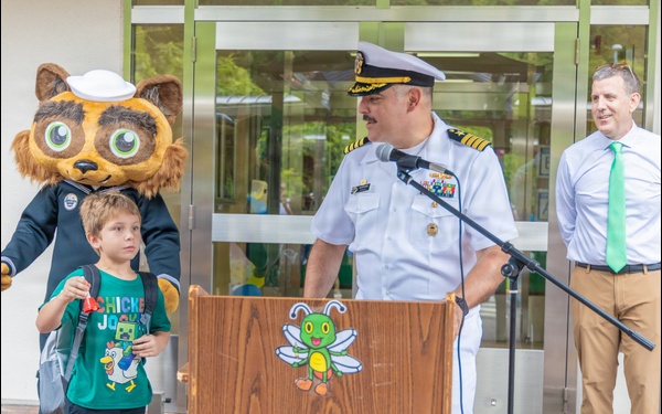 A naval officer in white dress uniform speaks at a podium during what appears to be a school event, likely a back-to-school ceremony. A young boy in a green 'Chicken Jockey' t-shirt stands beside a bear mascot wearing a sailor hat, while a man in a dress shirt and green tie looks on. The podium features a cartoon bee mascot logo, suggesting a school or educational institution setting.