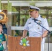 A naval officer in white dress uniform speaks at a podium during what appears to be a school event, likely a back-to-school ceremony. A young boy in a green 'Chicken Jockey' t-shirt stands beside a bear mascot wearing a sailor hat, while a man in a dress shirt and green tie looks on. The podium features a cartoon bee mascot logo, suggesting a school or educational institution setting.