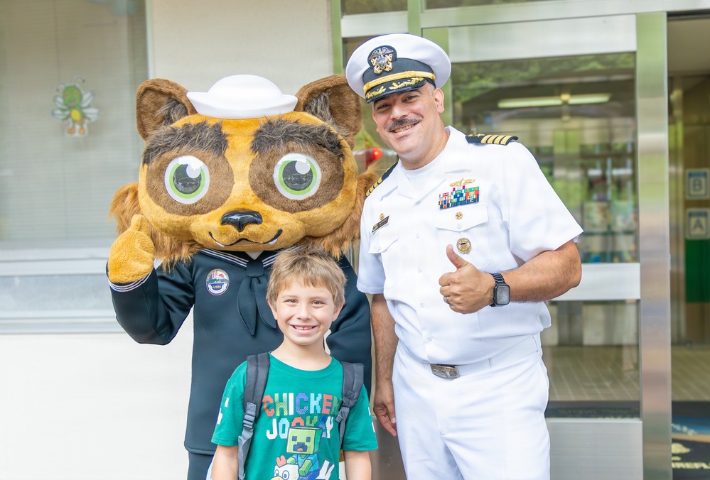 A U.S. Navy officer in white dress uniform poses with a young child and a costumed animal mascot wearing a sailor hat and navy uniform. The officer is giving a thumbs-up gesture while the child smiles broadly at the camera. The setting appears to be outside a building, likely during a community or school outreach event.