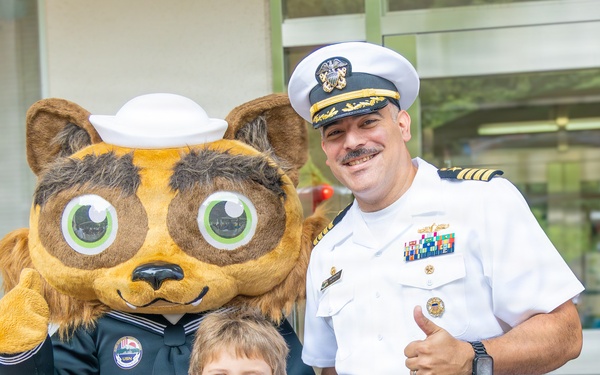 A U.S. Navy officer in white dress uniform poses with a young child and a costumed animal mascot wearing a sailor hat and navy uniform. The officer is giving a thumbs-up gesture while the child smiles broadly at the camera. The setting appears to be outside a building, likely during a community or school outreach event.