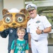 A U.S. Navy officer in white dress uniform poses with a young child and a costumed animal mascot wearing a sailor hat and navy uniform. The officer is giving a thumbs-up gesture while the child smiles broadly at the camera. The setting appears to be outside a building, likely during a community or school outreach event.