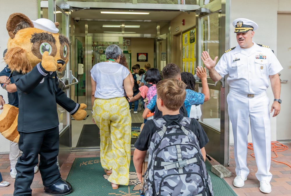 A naval officer in white dress uniform and a bear mascot dressed in a Navy uniform are greeting children at the entrance of what appears to be a school. The officer is giving a high-five to a student as children and an adult woman enter the building. The scene suggests a back-to-school or community outreach event involving the U.S. Navy.
