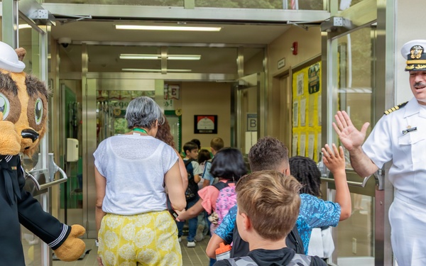 A naval officer in white dress uniform and a bear mascot dressed in a Navy uniform are greeting children at the entrance of what appears to be a school. The officer is giving a high-five to a student as children and an adult woman enter the building. The scene suggests a back-to-school or community outreach event involving the U.S. Navy.