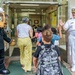 A naval officer in white dress uniform and a bear mascot dressed in a Navy uniform are greeting children at the entrance of what appears to be a school. The officer is giving a high-five to a student as children and an adult woman enter the building. The scene suggests a back-to-school or community outreach event involving the U.S. Navy.