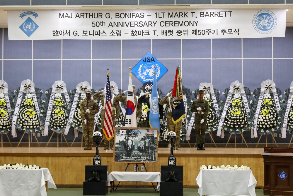 This image depicts the 50th Anniversary Ceremony honoring Major Arthur G. Bonifas and 1st Lieutenant Mark T. Barrett, as indicated by the banner at the top. The ceremony appears to be held at a Joint Security Area (JSA) venue, featuring military honor guards, national flags, and memorial floral wreaths. A memorial photograph of the two officers is displayed on an easel at the center of the stage.