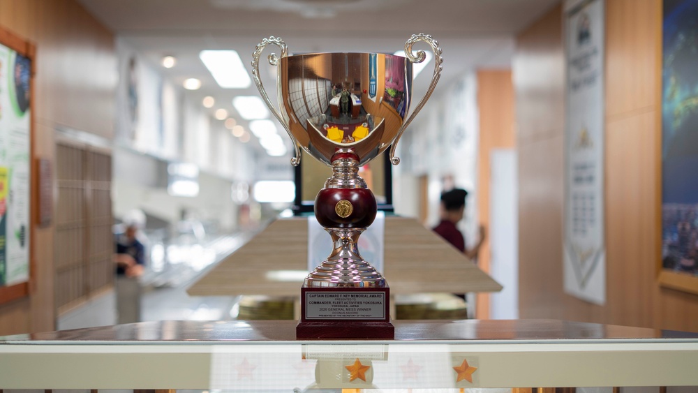 A large silver trophy cup with ornate handles and a dark red/maroon stem is displayed on a glass surface in what appears to be a military or institutional building. The trophy bears a plaque that references the 'Captain Edward F. Ney Memorial Award' and mentions 'Commander, Fleet Activities Yokosuka, Japan.' The background shows a blurred corridor with people and bulletin boards.